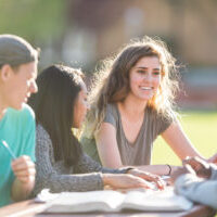 A coed, multiethnic group of college students study on center campus lawn on a beautiful spring day. They are sitting at a picnic table and are reading through their textbooks. The focus is on a Caucasian female who is smiling at one of her classmates.